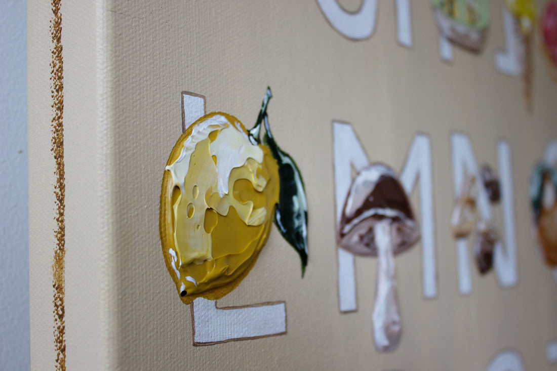 Alfabeto original painting by Marissa Lico. Close-up of the alphabet with a painted L and a yellow lemon on a beige background.