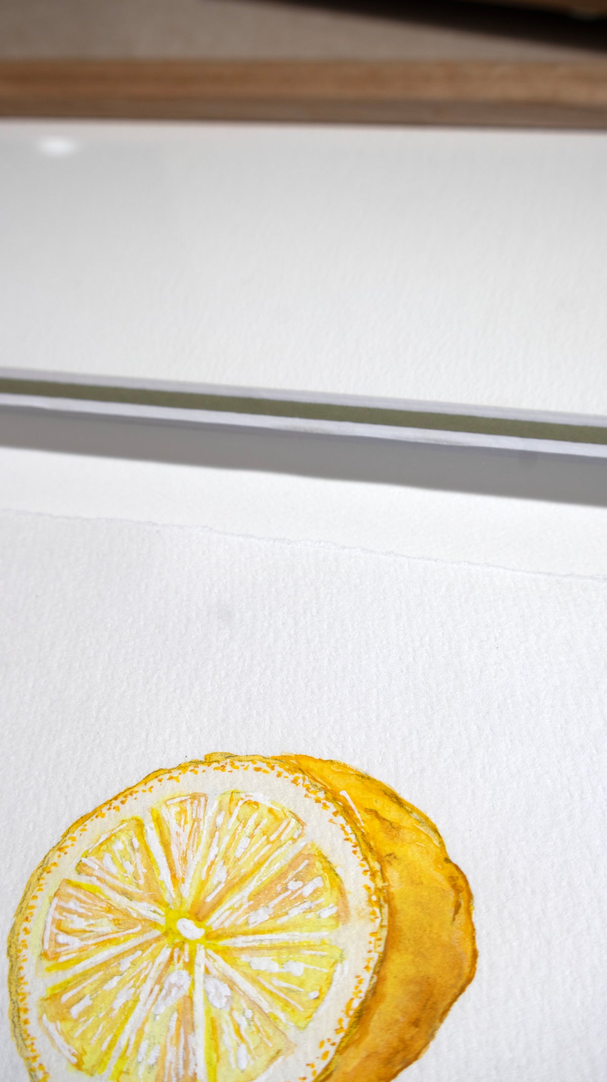 A close-up of a watercolour painting of a lemon slice on textured white paper, showing yellow tones with a green accent matt board and wooden oak frame in the background.