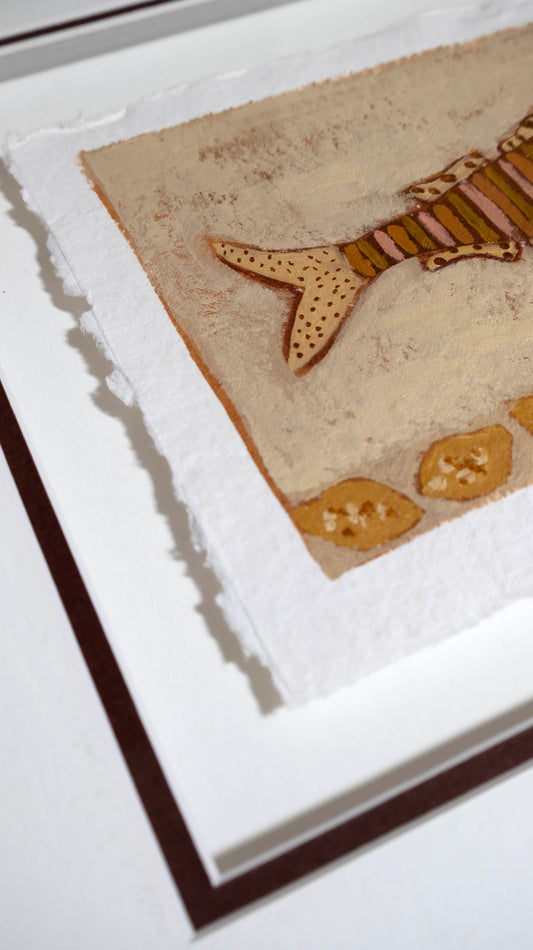 A close-up of framed wall art showing a textured beige background with a stylised fish motif in tan, brown, and muted orange, plus small decorative shapes near the bottom.