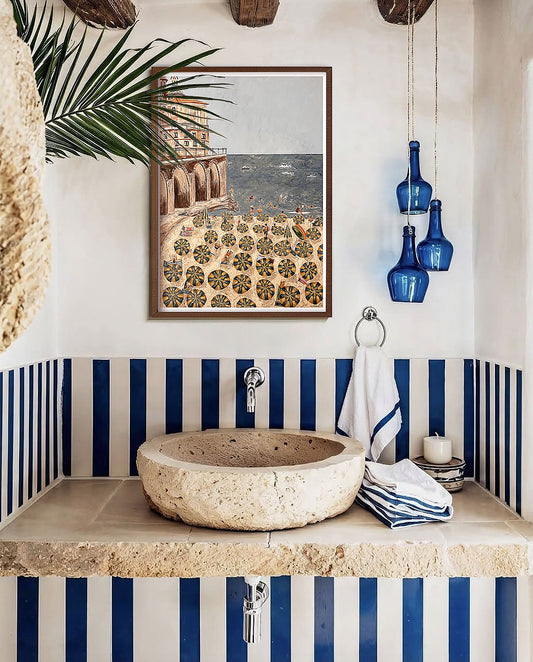 A framed art print depicting a Mediterranean beach scene with a patterned plaza, styled in an Italian bathroom setting above a stone basin sink with blue and white striped accents.