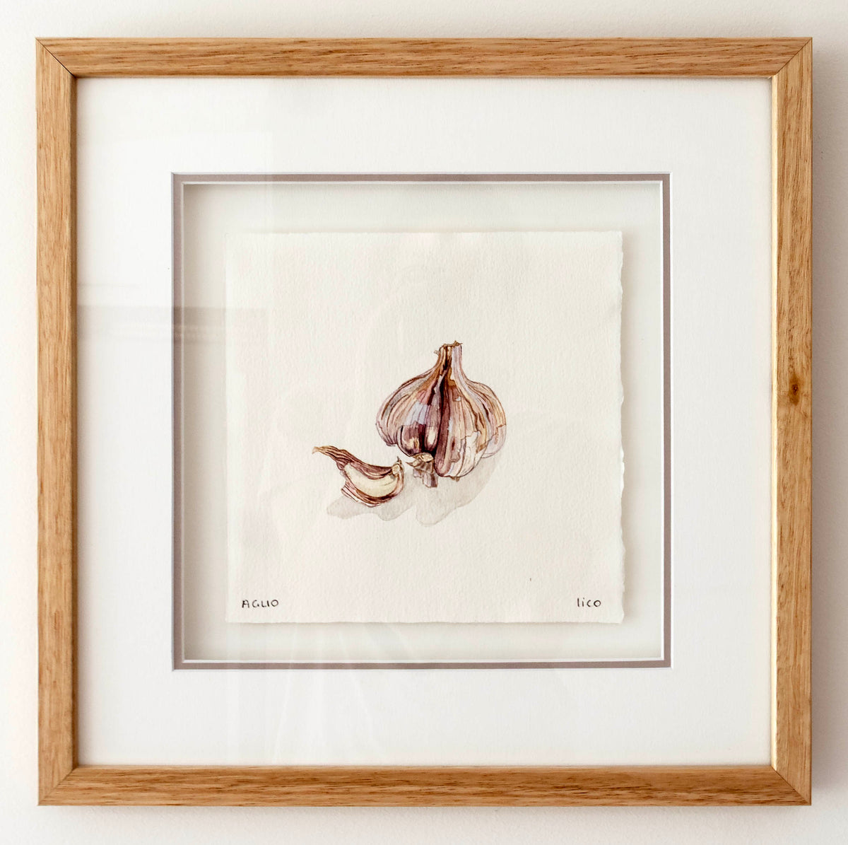 A framed watercolour painting of garlic bulbs and cloves on textured white paper, matted with a wide white border and enclosed in a light oak wood frame.