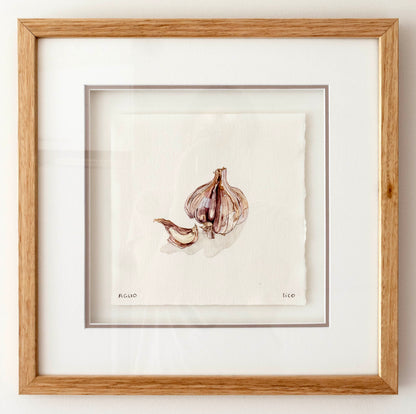 A framed watercolour painting of garlic bulbs and cloves on textured white paper, matted with a wide white border and enclosed in a light oak wood frame.