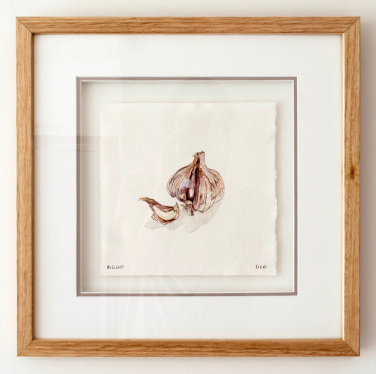 A framed watercolour painting of garlic bulbs and cloves on textured white paper, matted with a wide white border and enclosed in a light oak wood frame.