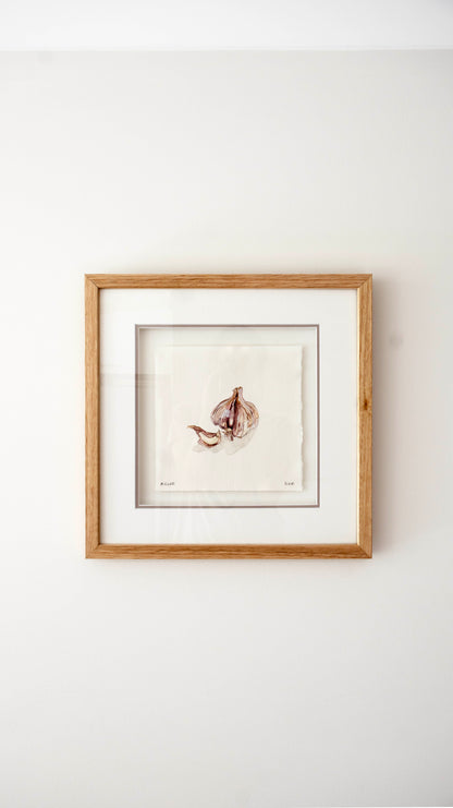 A square framed art print featuring a delicate watercolour illustration of garlic bulbs and stems, set against a white mat and a light natural wood frame, mounted on a plain white wall.