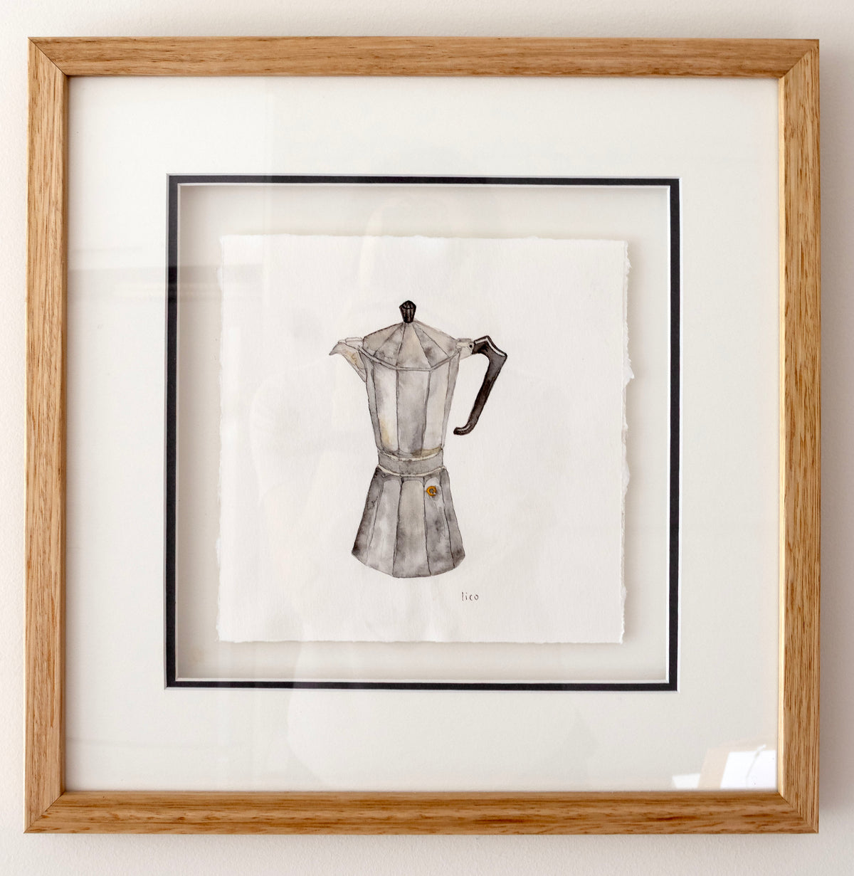 Framed watercolour artwork of a classic moka pot, featuring a detailed watercolour illustration of the coffee maker on textured white paper, set within a navy inner mat frame and a light oak wood outer frame.