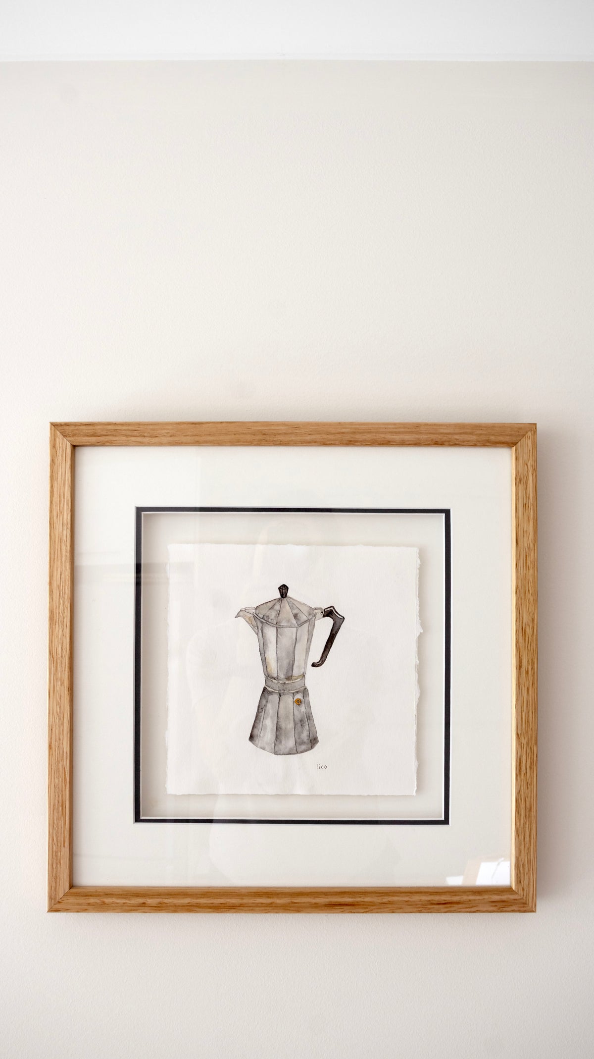 A framed artwork featuring a black and white watercolor illustration of a moka pot, displayed on a light beige wall, by artist Marissa Lico.