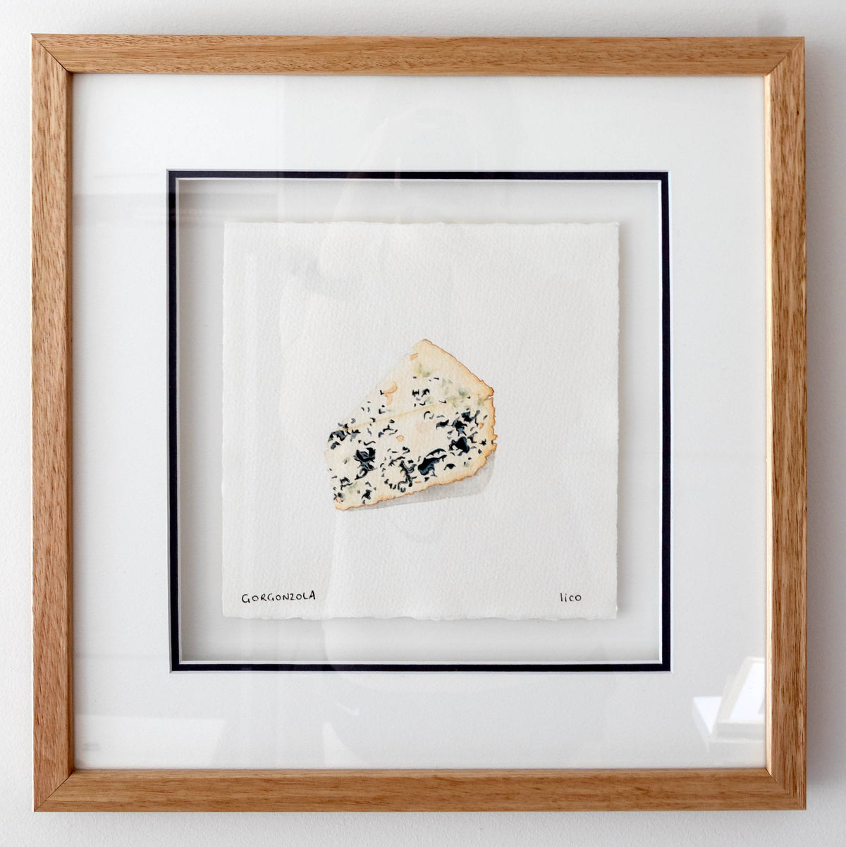 Framed artwork featuring a single slice of blue cheese on a white background, enclosed in a light oak wood frame with a white mat and navy inner border.