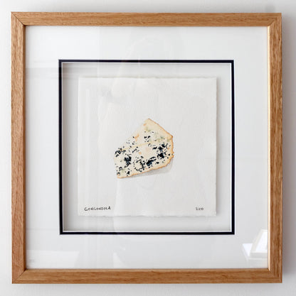 Framed artwork featuring a single slice of blue cheese on a white background, enclosed in a light oak wood frame with a white mat and navy inner border.