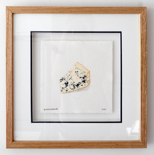 Framed artwork featuring a single slice of blue cheese on a white background, enclosed in a light oak wood frame with a white mat and navy inner border.