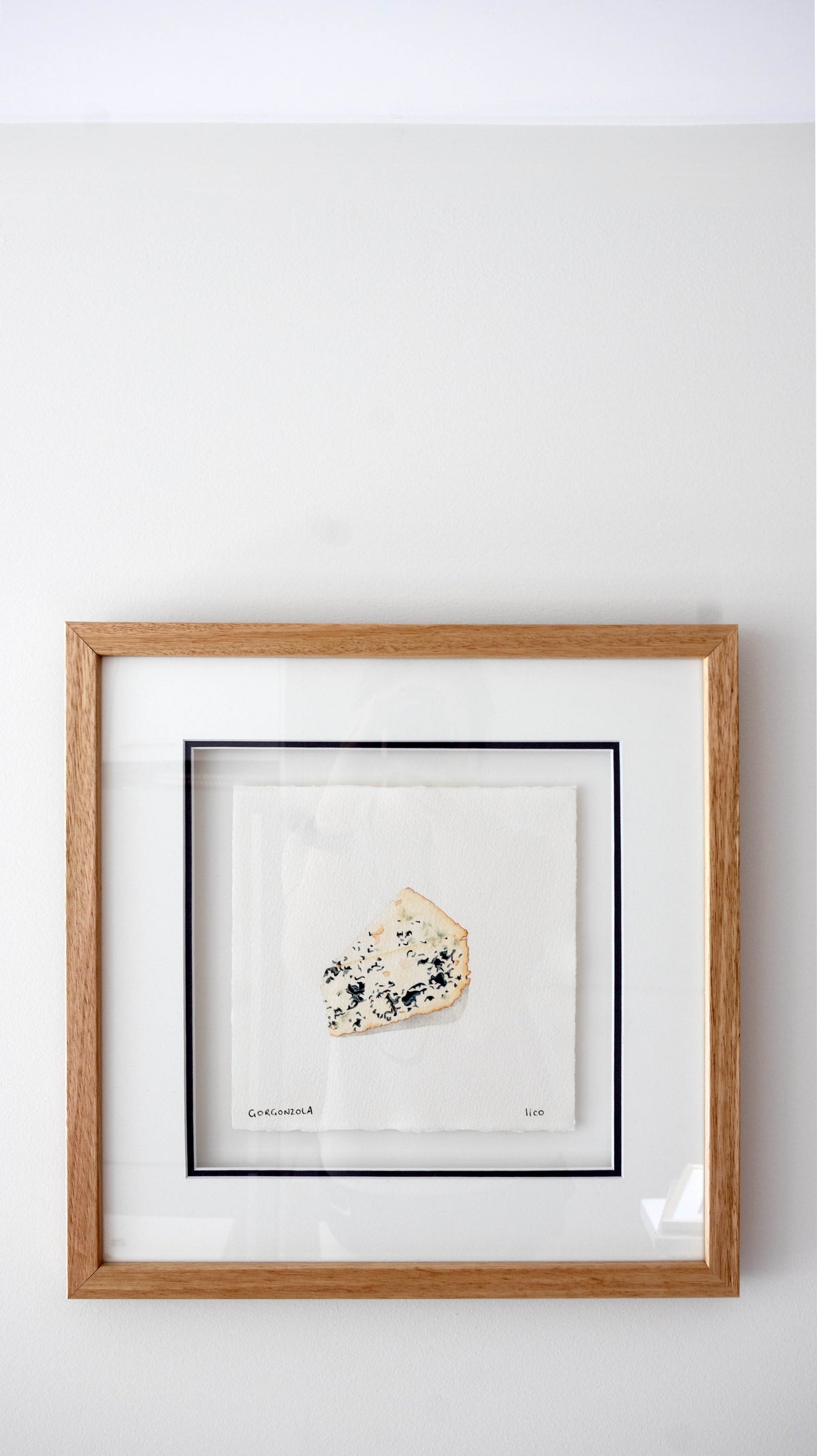 Framed wall art featuring a small gorgonzola blue cheese wedge mounted on a white mat, set in a light wood frame against a white wall.