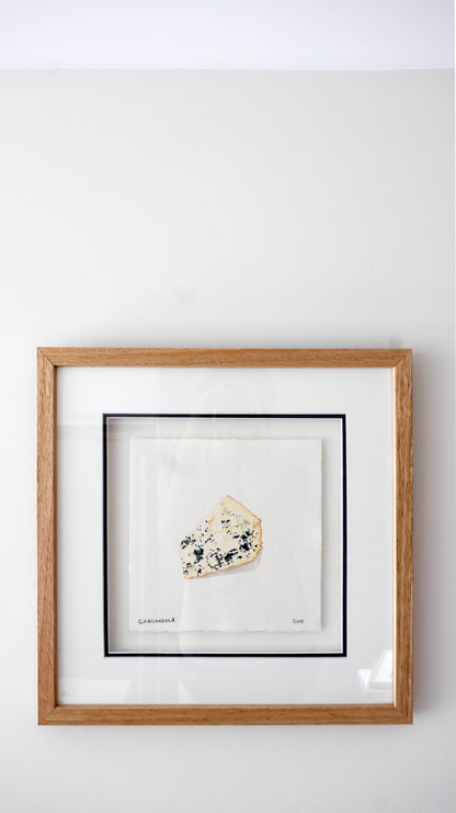 Framed wall art featuring a small gorgonzola blue cheese wedge mounted on a white mat, set in a light wood frame against a white wall.