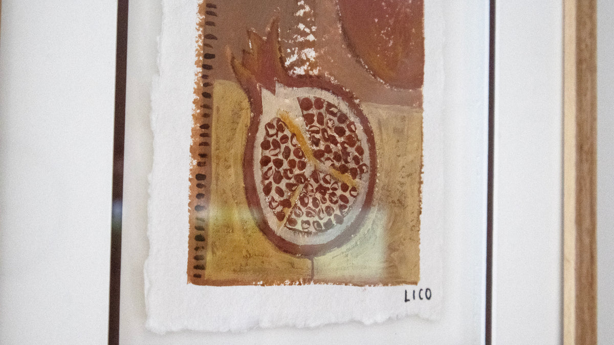 A framed Italian artwork featuring a pomegranate painting in earthy brown, tan, and white tones, mounted on a white wall with a light wood frame visible at the right edge, by Marissa Lico.