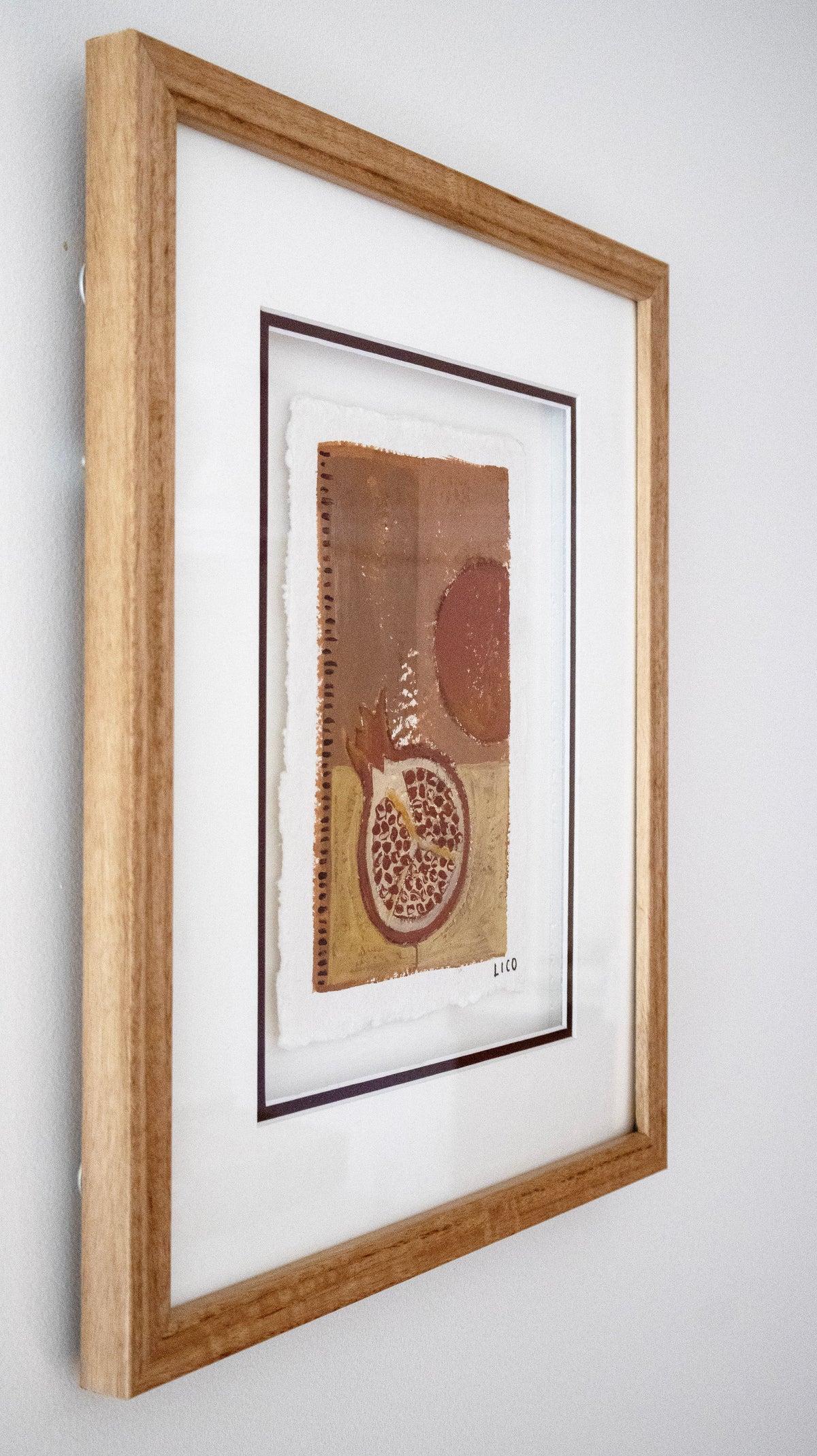 A framed original painting by Marissa Lico featuring earthy brown, tan, and terracotta tones with a pomegranate motif, mounted in a light oak wood frame with a white mat against a neutral light neutral wall.