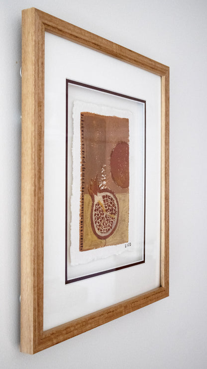 A framed original painting by Marissa Lico featuring earthy brown, tan, and terracotta tones with a pomegranate motif, mounted in a light oak wood frame with a white mat against a neutral light neutral wall.
