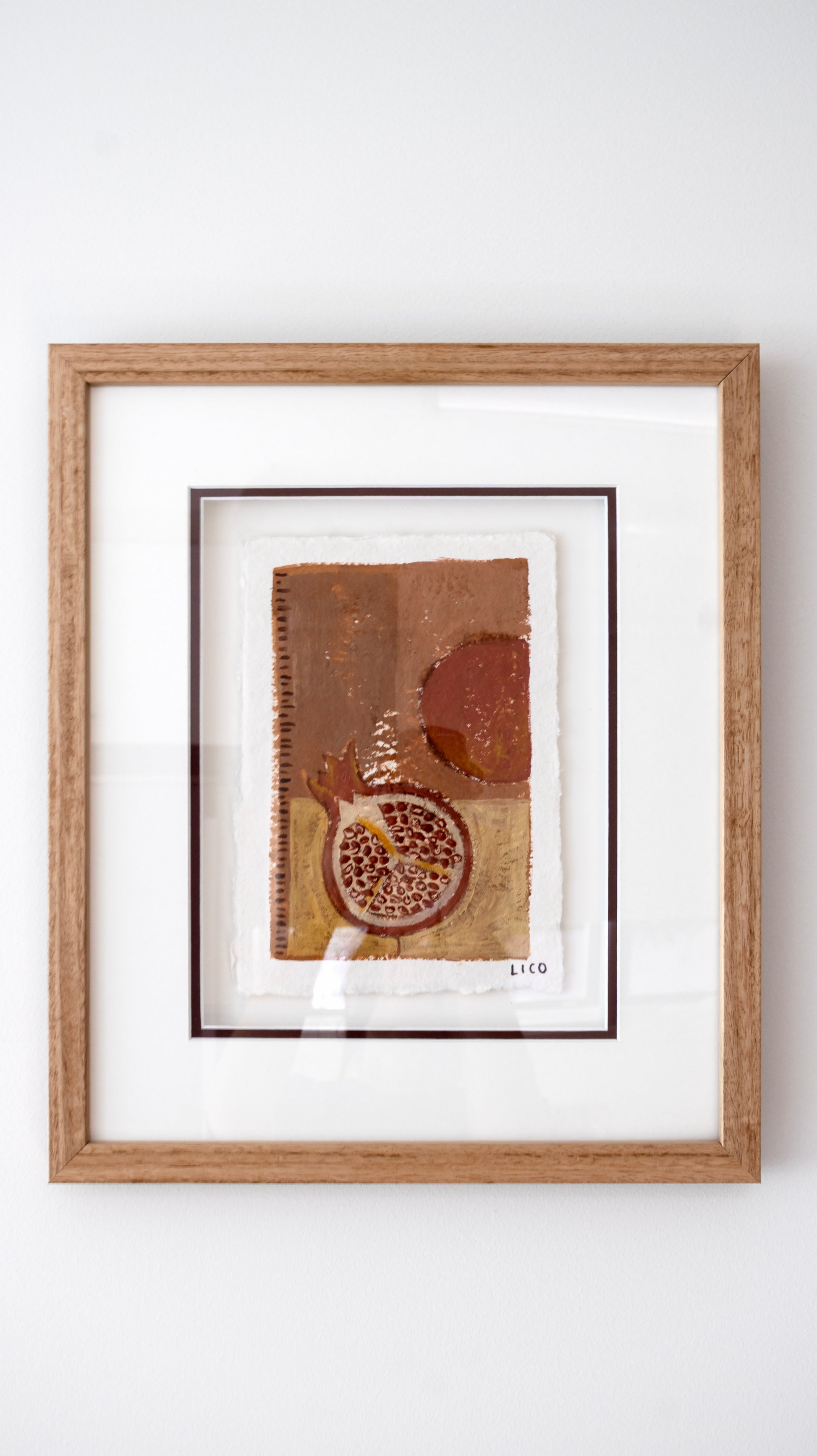 A framed Italian abstract artwork featuring textured brown, beige, and terracotta tones with a pomegranate motif, presented in a light wood oak frame with a white mat against a white wall.