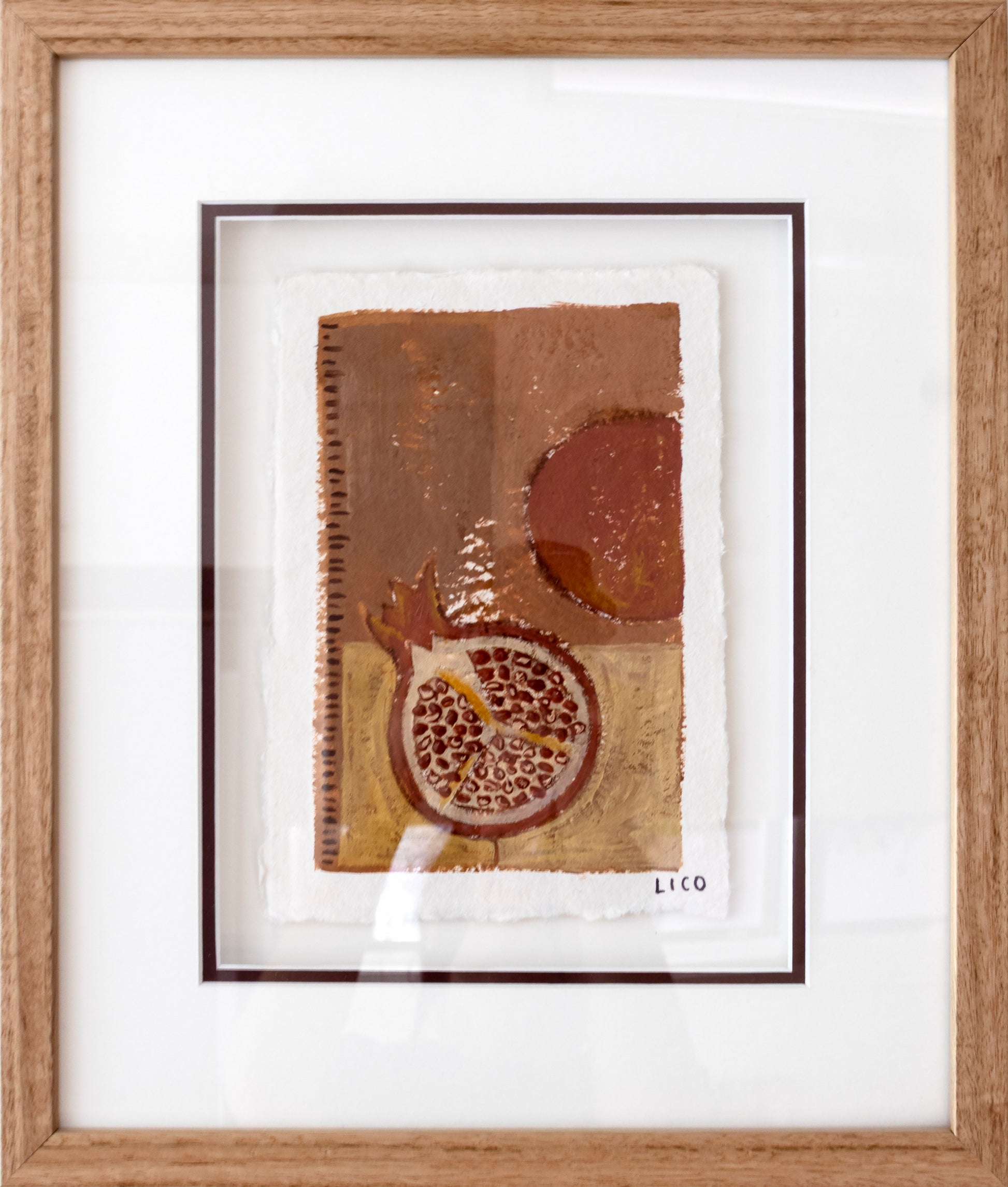 A framed abstract still life artwork featuring a pomegranate and warm earthy tones, presented in a light oak wood frame with a white mat.