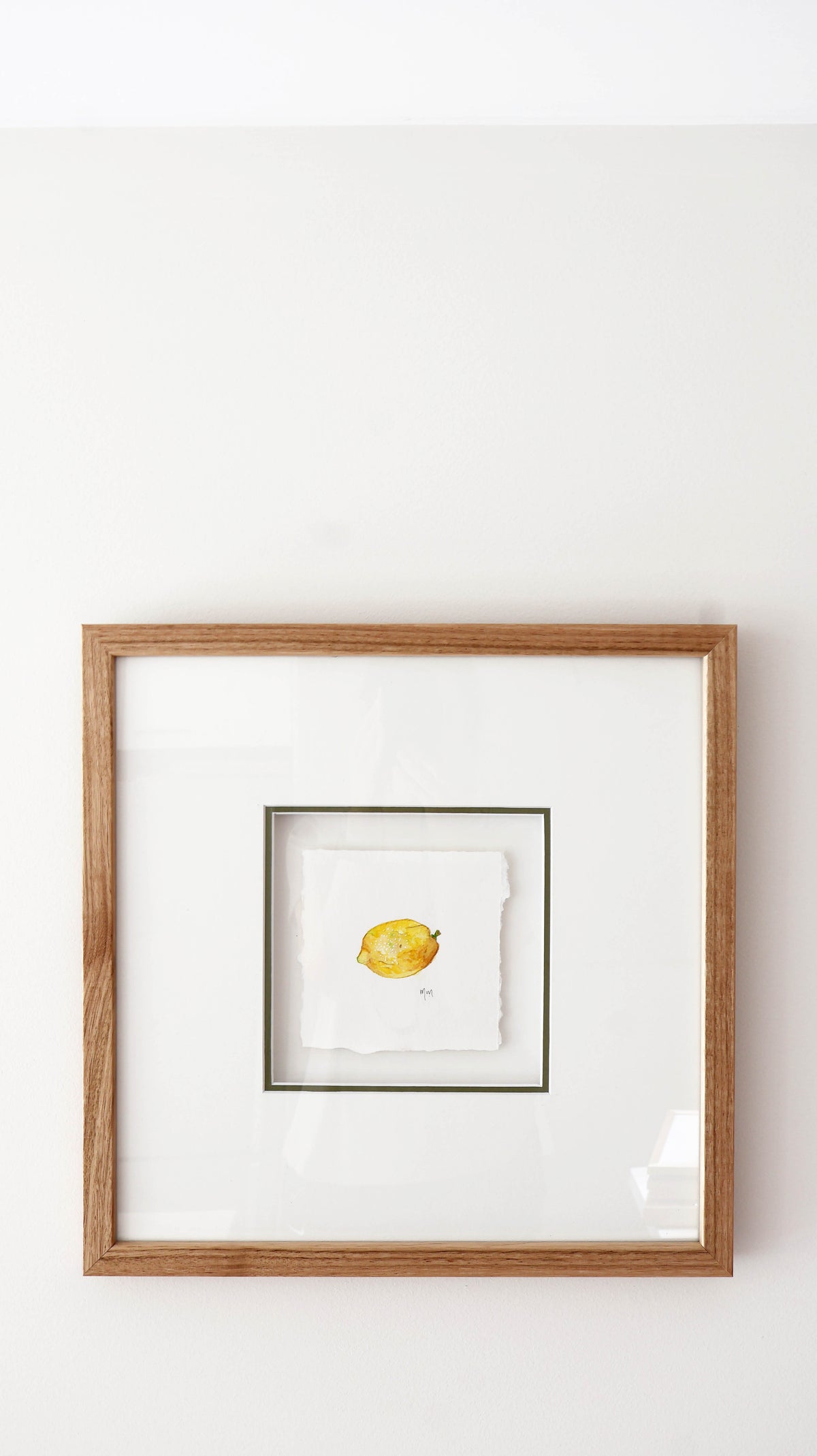 A square framed artwork featuring a small yellow watercolour lemon on layered white paper, set within a white mat and a light brown oak wood frame against a white wall.