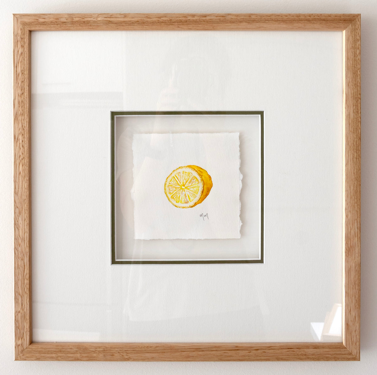 Framed square artwork featuring a small yellow lemon watercolour illustration on white textured paper, set within a white mat and a light natural wood oak frame.