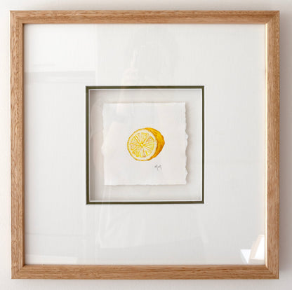 Framed square artwork featuring a small yellow lemon watercolour illustration on white textured paper, set within a white mat and a light natural wood oak frame.