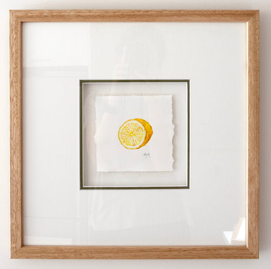 Framed square artwork featuring a small yellow lemon watercolour illustration on white textured paper, set within a white mat and a light natural wood oak frame.