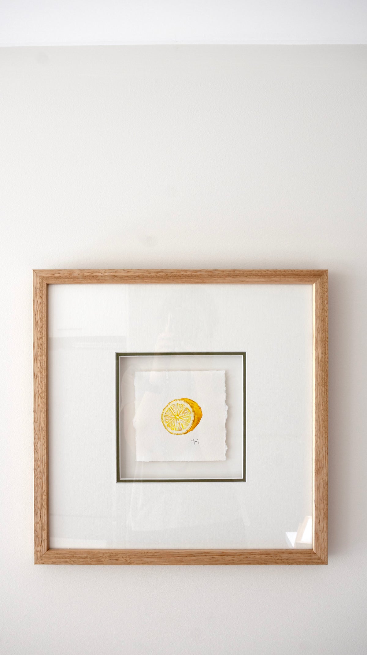 A square framed artwork featuring a yellow lemon slice illustration on a white background, set in a light natural oak wood frame with a white mat against a white wall.