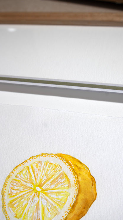 A close-up of a watercolour painting of a lemon slice on textured white paper, showing yellow tones with a green accent matt board and wooden oak frame in the background.