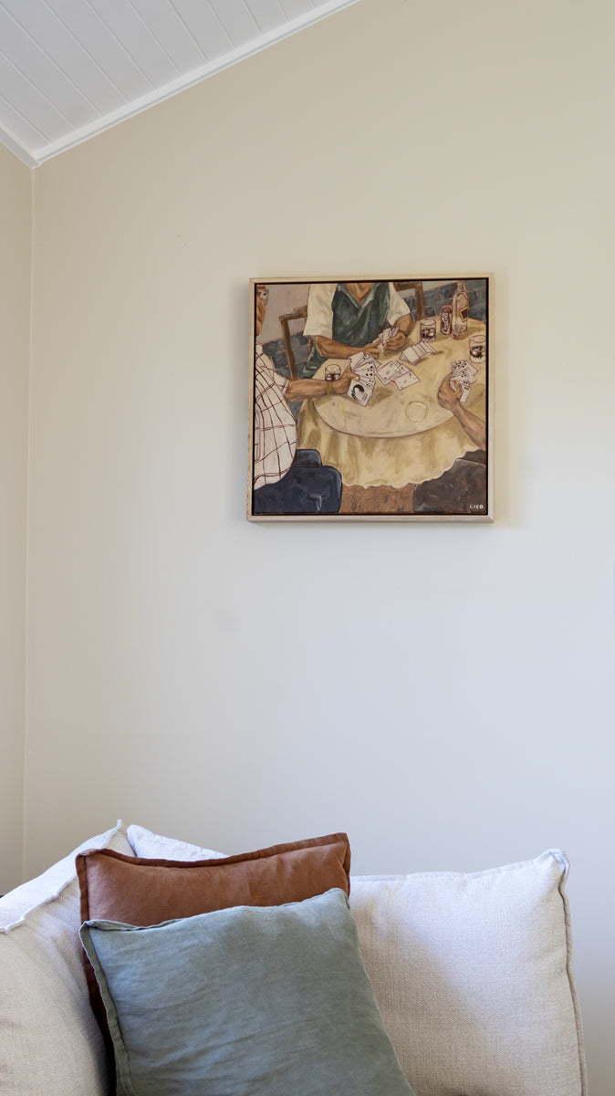 Original Italian-inspired painting of a family card game hung above a linen couch in a warm neutral living room setting.