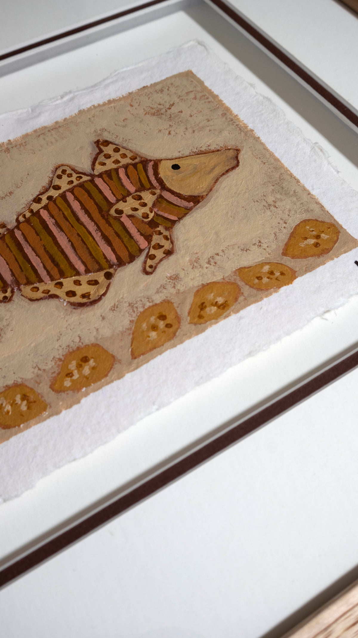 A framed handmade art piece featuring a painted fish and lemons on a textured beige background, photographed at an angle.