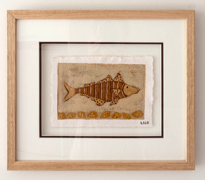 Framed folk artwork featuring a brown striped fish on a textured beige background, matted with white and housed in a light natural wood frame.