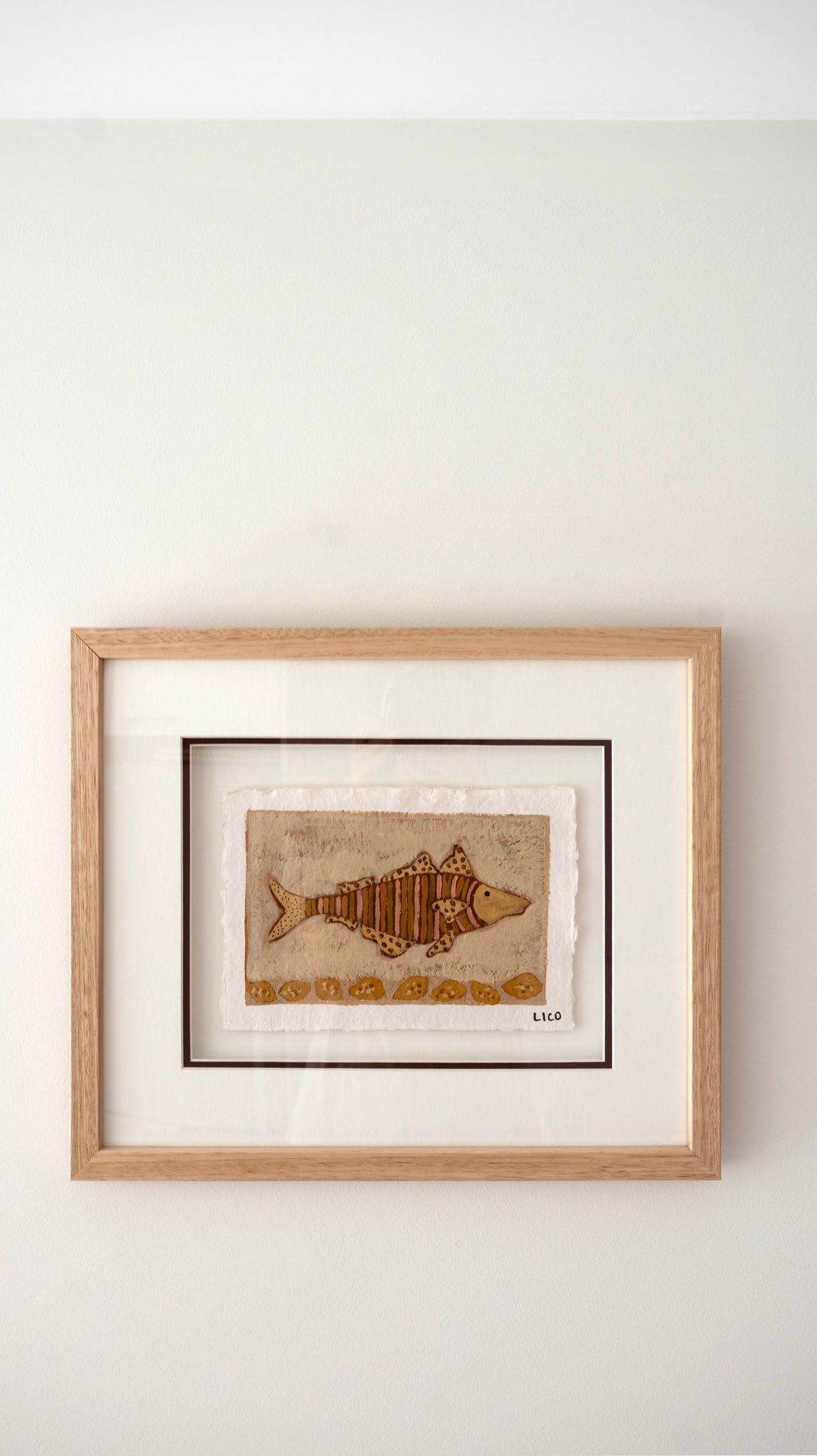 Framed wall art featuring a painted fish in warm brown and gold tones on a textured beige background, mounted with a white mat and light natural oak wood frame against a plain off-white wall.