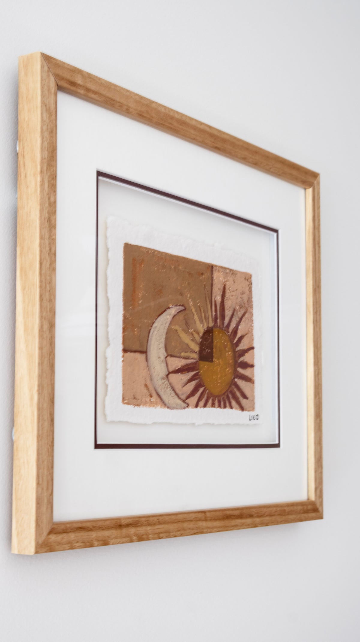 Framed abstract art painting original featuring a sun and crescent moon design in earthy brown, tan, and gold tones, mounted in a light natural oak wood frame with a white mat against a white wall.