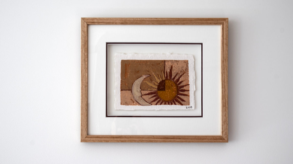 A framed original painting featuring a textured sun and crescent moon design in earthy brown, tan, and gold tones, set within a white mat and a light wood frame against a plain white wall.