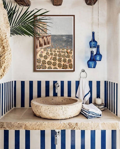 A framed art print depicting a Mediterranean beach scene with a patterned plaza, styled in an Italian bathroom setting above a stone basin sink with blue and white striped accents.